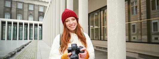 sorridente testa Rossa ragazza fotografo, assunzione immagini nel città, fa fotografie all'aperto su professionale telecamera