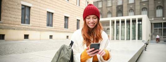 giovane testa Rossa donna con smartphone, seduta all'aperto con zaino, alunno guardare a sua mobile Telefono, testi Messaggio foto