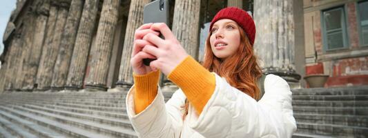 ritratto di giovane testa Rossa donna prende fotografie su sua mobile Telefono telecamera, utilizzando smartphone per catturare bene tiro all'aperto