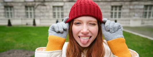 ritratto di divertente e carino testa Rossa ragazza mette su rosso cappello, Spettacoli lingua e strizza l'occhio a telecamera, sorrisi felicemente, gode grande tempo metereologico nel parco foto