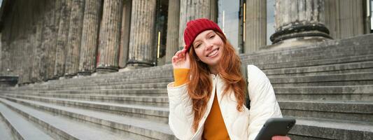 ritratto di giovane testa Rossa donna nel rosso cappello, seduta su scale, turista sembra a sua mobile Telefono, riposa su scala di Museo, si connette pubblico Wi-Fi foto