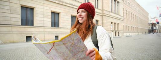 bellissimo testa Rossa donna, turista con città carta geografica, esplora giro turistico storico punto di riferimento, a piedi in giro vecchio cittadina, sorridente felicemente foto