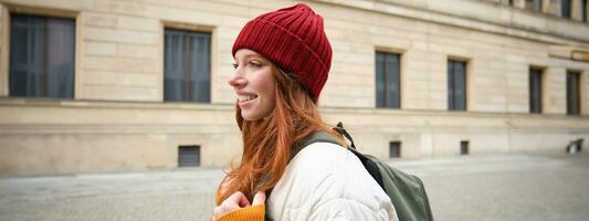 turismo e in viaggio. giovane testa Rossa donna sorridente, turista a piedi con zaino in giro città centro, giro turistico, detiene zaino con carta carta geografica foto