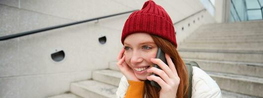 giovane elegante testa Rossa ragazza nel rosso cappello, si siede su strada e parla su mobile Telefono, ha telefono conversazione, anelli sua amico mentre rilassa all'aperto foto