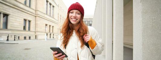 turismo e tecnologia. bellissimo testa Rossa ragazza turista, guardare per un' itinerario su applicazione, utilizzando città carta geografica su smartphone, lettura di giro turistico posti su mobile Telefono foto