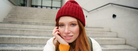 vicino su ritratto di bellissimo testa Rossa ragazza nel rosso cappello, urbano donna con lentiggini e Zenzero capelli, si siede su le scale su strada, sorrisi e sembra bellissima foto