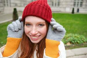 vicino su ritratto di bellissimo testa Rossa donna nel rosso a maglia cappello, caldo guanti, sorridente e guardare contento a telecamera, seduta nel parco foto