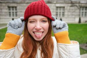 ritratto di divertente e carino testa Rossa ragazza mette su rosso cappello, Spettacoli lingua e strizza l'occhio a telecamera, sorrisi felicemente, gode grande tempo metereologico nel parco foto