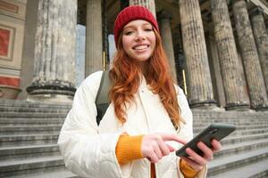 ritratto di giovane urbano ragazza con zaino, utilizzando smartphone, a piedi in giro città, guardare a carta geografica su mobile app, controllo direzione foto