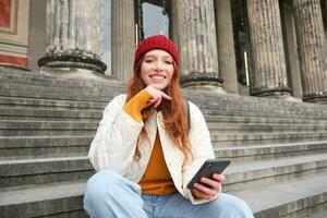 ritratto di giovane urbano ragazza nel rosso cappello, si siede su le scale vicino Museo, detiene mobile Telefono, si connette per pubblico Wi-Fi e surf rete, usi smartphone applicazioni foto