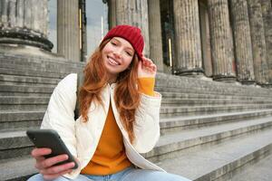 ritratto di giovane testa Rossa donna nel rosso cappello, seduta su scale, turista sembra a sua mobile Telefono, riposa su scala di Museo, si connette pubblico Wi-Fi foto