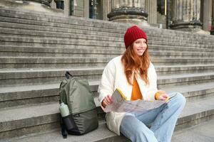 sorridente ragazza viaggiatore, turista si siede su le scale vicino Museo e detiene carta carta geografica, esplora città durante giro turistico avventura viaggio foto