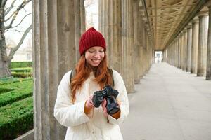 giovane testa Rossa femmina fotografo, fa stile di vita tiro nel città centro, prende fotografie e sorrisi, sembra per Perfetto sparo, fa immagine