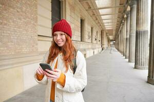 sorridente testa Rossa ragazza nel rosso cappello, sembra a carta geografica su smartphone app, controlli sua Posizione, ricerche per un' ostello. turista ricerche per popolare attrazioni su mobile Telefono foto