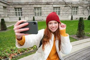 bellissimo sorridente Zenzero ragazza nel cappello, in posa per foto su mobile Telefono, prende autoscatto nel parco con carino viso espressione