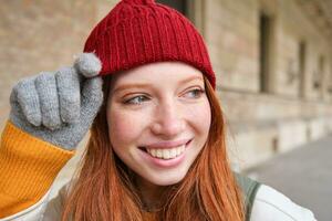 colpo alla testa di contento testa Rossa ragazza con lentiggini, indossa rosso cappello e guanti nel inverno, passeggiate in giro città su freddo tempo metereologico e sorrisi foto