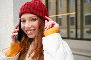 giovane persone e mobile connessione. contento testa Rossa ragazza parla su Telefono, fa telefono chiamata, sta all'aperto con zaino e usi smartphone App foto