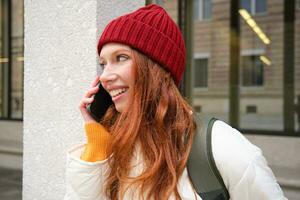 contento bellissimo ragazza, testa Rossa turista parla su mobile Telefono, fabbricazione un' chiamata all'estero, ha conversazione su smartphone, in piedi con zaino all'aperto foto