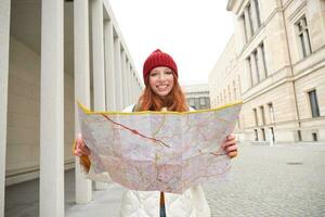 bellissimo testa Rossa donna, turista con città carta geografica, esplora giro turistico storico punto di riferimento, a piedi in giro vecchio cittadina, sorridente felicemente foto