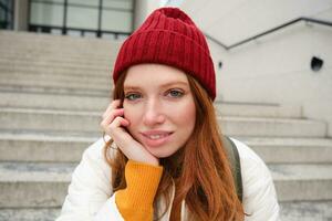 vicino su ritratto di bellissimo testa Rossa ragazza nel rosso cappello, urbano donna con lentiggini e Zenzero capelli, si siede su le scale su strada, sorrisi e sembra bellissima foto