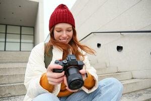 urbano persone e stile di vita. contento testa Rossa donna prende fotografie, Tenere professionale digitale telecamera, Fotografare su strade foto