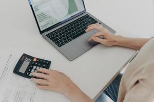 La donna irriconoscibile si siede alla scrivania con i documenti della calcolatrice del computer portatile calcola le spese mensili