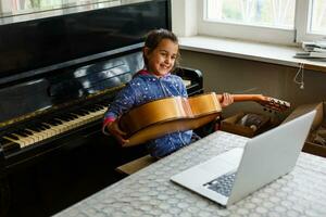 contento poco bambino nel denim camicia apprendimento per giocare chitarra mentre Guardando Lezioni a il computer portatile e sorridente a casa foto