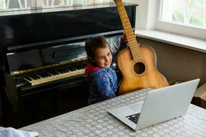 contento poco bambino nel denim camicia apprendimento per giocare chitarra mentre Guardando Lezioni a il computer portatile e sorridente a casa foto