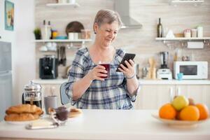 tempo libero mattina per anziano donna utilizzando moderno tecnologia nel luminosa accogliente cucina. autentico anziano persona utilizzando moderno smartphone Internet tecnologia. in linea comunicazione collegato per il mondo, anziano tempo libero tempo con aggeggio a la pensione età foto