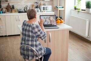 anziano donna agitando per sua figlia. contento anziano donna durante un' video conferenza con famiglia utilizzando il computer portatile nel cucina. in linea chiamata con figlia e nipote. anziano persona utilizzando moderno comunicazione in linea Internet ragnatela tecnologia. foto