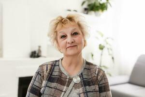 sorridente mezzo anziano maturo grigio dai capelli donna guardare a telecamera, contento vecchio signora in posa a casa interno, positivo singolo anziano pensionato femmina seduta su divano nel vivente camera colpo alla testa ritratto foto