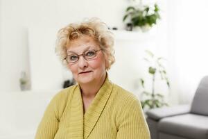 sorridente mezzo anziano maturo grigio dai capelli donna guardare a telecamera, contento vecchio signora nel bicchieri in posa a casa interno, positivo singolo anziano pensionato femmina seduta su divano nel vivente camera colpo alla testa ritratto foto