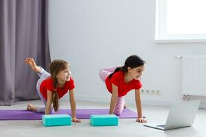 Due poco ragazze praticante yoga, allungamento, fitness di video su taccuino. lontano in linea formazione scolastica addestramento, aerobico a casa. salutare stile di vita, coronavirus, restare casa. bambini sport casa quarantena. foto