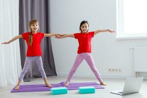 Due poco ragazze praticante yoga, allungamento, fitness di video su taccuino. lontano in linea formazione scolastica addestramento, aerobico a casa. salutare stile di vita, coronavirus, restare casa. bambini sport casa quarantena. foto