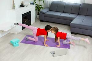 Due poco ragazze praticante yoga, allungamento, fitness di video su taccuino. lontano in linea formazione scolastica addestramento, aerobico a casa. salutare stile di vita, coronavirus, restare casa. bambini sport casa quarantena. foto