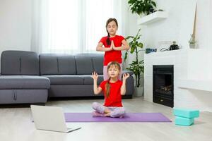 Due poco ragazze praticante yoga, allungamento, fitness di video su taccuino. lontano in linea formazione scolastica addestramento, aerobico a casa. salutare stile di vita, coronavirus, restare casa. bambini sport casa quarantena. foto