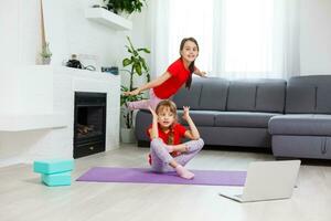 Due poco ragazze praticante yoga, allungamento, fitness di video su taccuino. lontano in linea formazione scolastica addestramento, aerobico a casa. salutare stile di vita, coronavirus, restare casa. bambini sport casa quarantena. foto