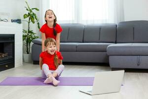 Due poco ragazze praticante yoga, allungamento, fitness di video su taccuino. lontano in linea formazione scolastica addestramento, aerobico a casa. salutare stile di vita, coronavirus, restare casa. bambini sport casa quarantena. foto