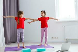 Due poco ragazze praticante yoga, allungamento, fitness di video su taccuino. lontano in linea formazione scolastica addestramento, aerobico a casa. salutare stile di vita, coronavirus, restare casa. bambini sport casa quarantena. foto