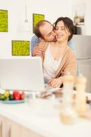 marito fabbricazione moglie Sorridi mentre baci sua nel cucina. coppia utilizzando il computer portatile. contento amorevole allegro romantico nel amore coppia a casa utilizzando moderno Wi-Fi senza fili Internet tecnologia foto