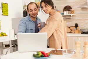 sorpreso moglie guardare a il computer portatile nel cucina con tazza di caffè nel mano. contento imprenditore marito. contento amorevole allegro romantico nel amore coppia a casa utilizzando moderno Wi-Fi senza fili Internet tecnologia foto