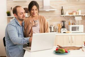 allegro imprenditore coppia utilizzando il computer portatile nel cucina. fidanzata Tenere tazza di caffè. contento amorevole allegro romantico nel amore coppia a casa utilizzando moderno Wi-Fi senza fili Internet tecnologia foto