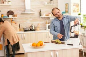 uomo miscelazione frutta nel miscelatore mentre moglie è guardare dentro frigorifero. salutare spensierato e allegro stile di vita, mangiare dieta e preparazione prima colazione nel accogliente soleggiato mattina foto