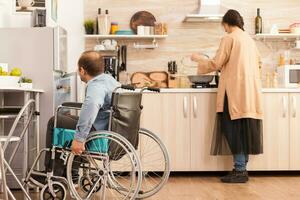 Disabilitato uomo nel sedia a rotelle si apre frigorifero mentre moglie è cucinando. Disabilitato paralizzato portatori di handicap uomo con a piedi invalidità integrando dopo un incidente. foto