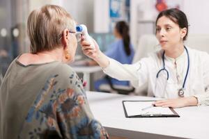 giovane medico utilizzando infrarosso termometro per prendere anziano donna temperatura nel ospedale ufficio. medico indossare bianca cappotto e stetoscopio e infermiera Lavorando su computer nel il sfondo. foto