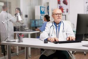 anziano uomo medico con grigio capelli Lavorando su computer mentre giovane collega è con un' paziente su ospedale corridoio e infermiera controllo analisi rapporto. foto