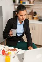 attività commerciale proprietario potabile caffè nel davanti di il computer portatile preparazione per incontro. concentrato attività commerciale donna nel il mattina multitasking nel il cucina prima andando per il ufficio, stressante modo di vita, carriera e obiettivi per incontrare. foto