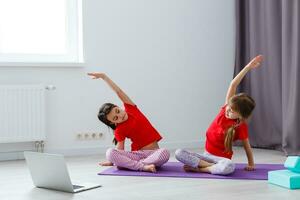 Due poco ragazze praticante yoga, allungamento, fitness di video su taccuino. lontano in linea formazione scolastica addestramento, aerobico a casa. salutare stile di vita, coronavirus, restare casa. bambini sport casa quarantena. foto