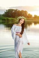 bellissimo nero dai capelli ragazza nel bianca Vintage ▾ vestito e ghirlanda di fiori in piedi nel acqua di lago. sole bagliore. foto
