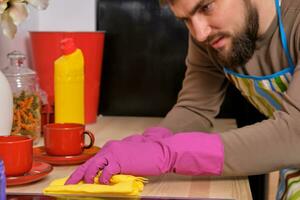 giovane bello barbuto uomo nel il cucina, indossare grembiule e rosa guanti pulisce il cucinando superficie utilizzando detersivi foto